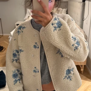 Teddyjacka med blå broderade blommor från Gina Tricot - Superfin beige teddyjacka från Gina Tricot med blå broderade blommor. Jackan har rund halsringning, knappar framtill och är långärmad. Aldrig använd med lapp kvar!💓💓