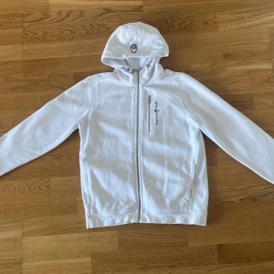 Vit hoodie från Sail Racing - Säljer en vit hoodie från Sail Racing med dragkedja framtill och logga på bröstet. Tröjan har huva och långa ärmar, perfekt för en sportig och avslappnad stil. Passar dig som gillar klassiska och stilrena plagg.