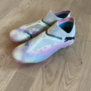 Puma fotbollsskor i vitt och rosa Elite - Säljer ett par vita och rosa fotbollsskor från Puma med stickad ovandel och högt skaft. Skorna har svarta Puma-loggor och sulan är i starkt rosa och vitt med fasta dobbar. Perfekta för dig som vill sticka ut på planen!