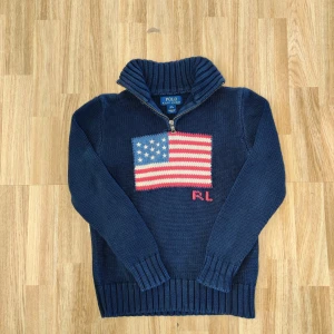 Marinblå stickad tröja från Polo Ralph Lauren - Säljer en marinblå stickad tröja från Polo Ralph Lauren med halv dragkedja och hög krage. Framsidan har en stor amerikansk flagga i rött, vitt och blått samt RL-broderi. Perfekt för kyliga dagar. M i kids