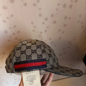 Gucci keps med monogram och röd-blå rand - Snygg keps från Gucci med klassiskt GG-monogram i beige och brunt. Kepsen har en röd och blå rand baktill och är tillverkad i bomullsmaterial. Perfekt accessoar för en trendig look. Helt ny. 