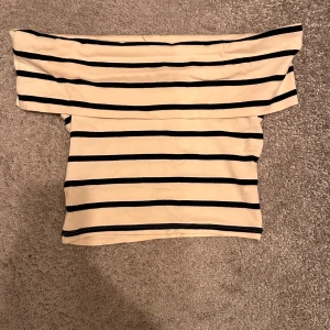 Offshoulder topp - Säljer en beige offshoulder topp med svarta horisontella ränder. Toppen har en bred krage som vilar över axlarna och är tillverkad i ett mjukt material. Perfekt för en trendig och avslappnad look.
