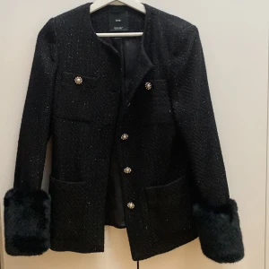 Svart kavaj med fuskpälsdetaljer från MNG AVTAGNINGSBARA PÄLSMUDDAR  - Säljer en svart kavaj från MNG med glittrig textur och dekorativa knappar framtill. Jackan har långa ärmar med breda fuskpälsdetaljer vid ärmsluten som är avtagningsbara(?!) och två stora fickor framtill. Perfekt för dig som vill sticka ut med stil. Nypris 1000kr använd ett tillfälle som ny. 