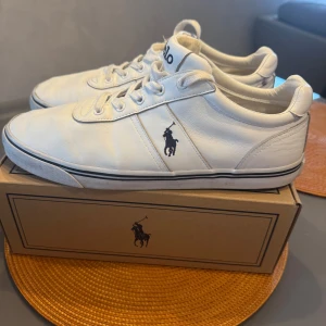 Vita sneakers från Polo Ralph Lauren - Säljer ett par klassiska vita sneakers från Polo Ralph Lauren med svart logga på sidan. Skorna har snörning, rund tå och platt sula. Perfekta till jeans eller shorts för en clean och stilren look.