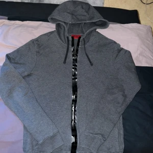 Grå mjukisbyxor och kofta från Hugo Boss - Säljer ett par grå mjukisbyxor och kofta från Hugo Boss med svarta detaljer vid fickorna och midjan. Byxorna har resår i midjan och benslut samt snörning för extra komfort. Perfekta för en avslappnad stil.