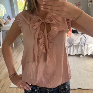 Rosa blus med volang och rosett från Fornarina - Supersöt ljusrosa blus från Fornarina med volangkantad v-ringning och en rosett! Ärmlös modell med rynkade axlar och glansigt material som ger en lyxig känsla😍