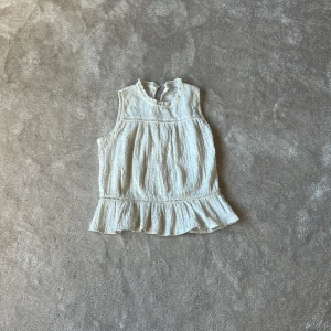 Vit ärmlös blus med spetsdetaljer från Zara - Säljer en söt vit ärmlös blus från Zara med broderade spetsdetaljer upptill och volang nertill. Blusen har en lätt skrynklig struktur och fina hålbroderier vid halsen. Perfekt för varma dagar!