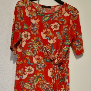 Röd blommig klänning från Betty Barclay - Färgstark röd klänning med blommigt mönster från Betty Barclay. Klänningen har korta ärmar, knyt i midjan och rund halsringning. Perfekt för dig som vill sticka ut med en livlig och somrig look.