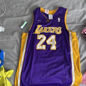 Lakers baskettröja Kobe Bryant #24 - Säljer en klassisk Los Angeles Lakers baskettröja med nummer 24 och Bryant på ryggen. Tröjan är lila med gula och vita detaljer, officiell NBA-logga och Adidas-märke. Perfekt för fans av Kobe Bryant och Lakers.