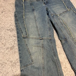 Blå jeansbyxor från Fashion Nova med patchwork-detaljer - Säljer ett par blå jeansbyxor från Fashion Nova med coola patchwork-sömmar och flera fickor. Byxorna har normal passform och stängs med dragkedja och knapp. Perfekta för dig som vill ha en unik streetstyle-look.