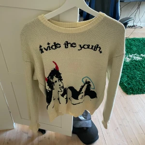 Beige stickad tröja från Divide The Youth - Säljer en beige stickad tröja från Divide The Youth med svart text och grafiskt motiv framtill. Tröjan har rund halsringning och långa ärmar. Perfekt för dig som vill ha något unikt och trendigt i garderoben.