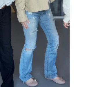 Ljusblå slitna bootcut jeans - Säljer ett par ljusblå bootcut jeans med slitningar och hål på knäna. Jeansen har klassisk femficksdesign och låg midja. Perfekta för en avslappnad och trendig look.