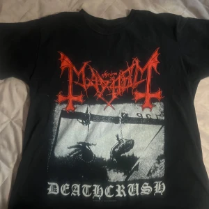 Svart band-t-shirt av Mayhem  - Svart t-shirt med stort tryck av Mayhem-loggan i rött och motiv från albumet Deathcrush i vitt. Klassisk passform och rund hals. Perfekt för dig som gillar metal och vill sticka ut med en ikonisk bandtröja.