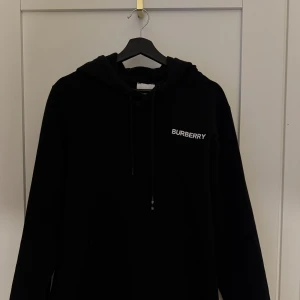 Svart hoodie från Burberry - Säljer en svart hoodie från Burberry med vit logotyp på bröstet. Tröjan har huva med dragsko och långa ärmar. Perfekt för en stilren och avslappnad look. Passar Medium & Large