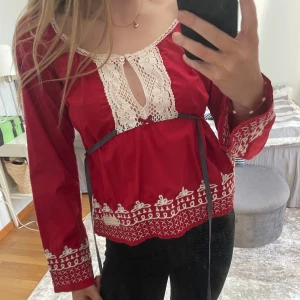 Röd blus med spetsdetaljer - Röd blus med vita spetsdetaljer och broderier längs ärmslut, nederkant och runt halsen. Blusen har lång ärm och en dekorativ öppning framtill vid halsen. Perfekt för dig som gillar bohemisk stil.