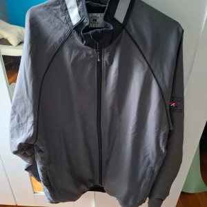 Grå vindjacka från Cutter & Buck - Säljer en grå vindjacka från Cutter & Buck med dragkedja framtill och broderad logga på ärmen. Jackan har lång ärm och reflekterande detaljer vid kragen. Perfekt för blåsiga dagar och enkel att matcha.