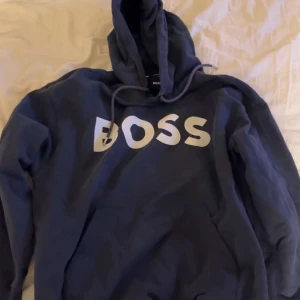 Mörkblå hoodie från hugo boss - Säljer en mörkblå hoodie från BOSS med stor vit logga på bröstet. Tröjan har huva med dragsko och en klassisk passform. Perfekt för en avslappnad stil.