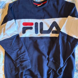 Blå sweatshirt från FILA - Säljer en blå sweatshirt från FILA med stor logga framtill. Tröjan har långa ärmar, ribbade muddar och en vit panel över bröstet och armarna. Perfekt för en sportig och avslappnad stil.
