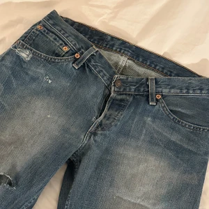 Blå slitna jeans Levis  - Säljer ett par klassiska blå Levis 501 jeans med slitningar och hål på benen. Jeansen har raka/bootcut ben, fem fickor och knappgylf. Perfekta för en avslappnad och trendig look.