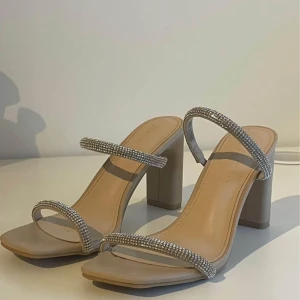 Beige sandaler med klack och glittriga remmar - Säljer ett par snygga beige sandaler med fyrkantig tå och bred klack. Skorna har två smala remmar täckta av glittrande stenar som ger en lyxig känsla. Perfekta för att lyfta vilken outfit som helst.
