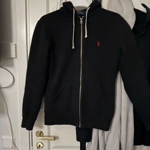 Svart hoodie från Polo Ralph Lauren - Svart hoodie från Polo Ralph Lauren med dragkedja och huva. I jättebra skick och jättefin. Klassisk röd logga broderad på bröstet och två fickor framtill. Perfekt för en avslappnad och stilren look.