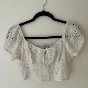 Vit broderad croppad blus från Hollister - Superfin vit croppad blus från Hollister med broderade hålmönster och puffärmar. Toppen har knyt framtill och är tillverkad i bomull. Perfekt för sommardagar och ger en romantisk vibe.