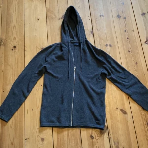 Mörkgrå hoodie med dragkedja från Jack & Jones - Mörkgrå hoodie från Jack & Jones i mjukt material. Tröjan har dragkedja framtill, huva med snören och långa ärmar. Perfekt för en avslappnad stil och enkel att matcha med andra plagg.