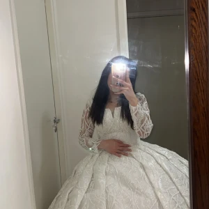 Vit långklänning med spetsdetaljer - Säljer en fantastisk vit långklänning med vackra spetsdetaljer och lång ärm. Klänningen har en voluminös kjol och elegant mönster över hela tyget, perfekt för speciella tillfällen.