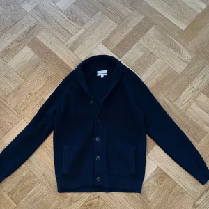 Mörkblå kofta från M&S - Säljer en klassisk mörkblå kofta från M&S med knappar framtill och två fickor. Koftan har lång ärm och är stickad i ett mjukt material, perfekt för lager-på-lager. Snygg och enkel att matcha till olika outfits.