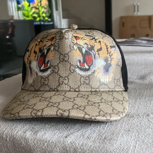 Gucci keps med tigertryck - Snygg keps från Gucci med klassiskt GG-mönster i beige och två stora tigerhuvuden framtill. Baksidan är i svart mesh och justerbar med grön-röd randig rem. Perfekt statement-piece för dig som vill sticka ut.