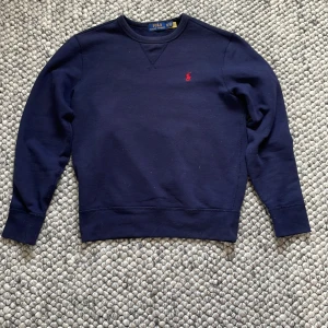 Mörkblå sweatshirt från Polo Ralph Lauren - Säljer en klassisk mörkblå sweatshirt från Polo Ralph Lauren med röd broderad logga på bröstet. Tröjan har rund halsringning och långa ärmar. Säljes då den inte passar längre.