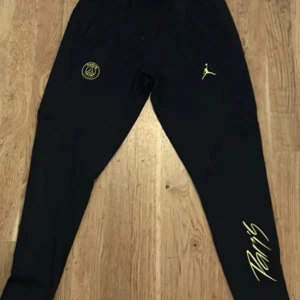 Svarta träningsbyxor PSG x Jordan - Svarta träningsbyxor med PSG-logga och Jordan-logga i gult. Byxorna har smal passform och resår i midjan. Snygg Paris-text på ena benet. Perfekta för dig som gillar sportig stil.