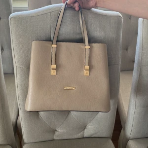 Beige handväska - Snygg beige handväska med guldfärgade detaljer och dubbla handtag. Väskan har en stilren och klassisk design, perfekt för att lyfta vilken outfit som helst. Tillverkad i syntetmaterial med struktur.