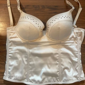 korsett-topp med detaljer - Säljer en beige/guldig korsett-topp med vadderade kupor och små glittriga stenar upptill. Justerbara axelband och hyskknäppning bak. Snygg och figurnära modell i glansigt material. 