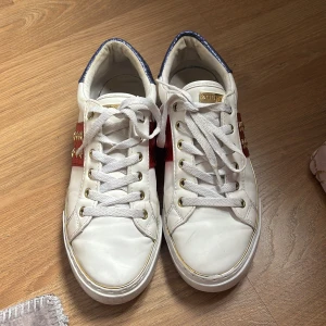 Vita sneakers från Guess med stjärndetaljer - Säljer ett par vita sneakers från Guess med röda paneler och guldstjärnor på sidorna. Skorna har blå hälkappa med reptilmönster, vita snören och guldfärgade detaljer vid snörningen. Perfekta för dig som vill sticka ut med snygga detaljer.