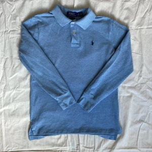 Ljusblå långärmad pikétröja från Polo Ralph Lauren - Säljer en klassisk ljusblå långärmad pikétröja från Polo Ralph Lauren med krage och knappar framtill. Tröjan har broderad logga på bröstet och är tillverkad i mjuk bomull. Perfekt för en stilren och avslappnad look. Tröjan är M i barnstorlek, för 8-9 åringar. 140-150cm