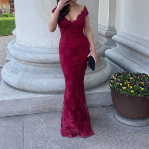 Vinröd långklänning med spetsdetaljer  - Säljer en elegant vinröd långklänning med v-ringning och spetsdetaljer. Klänningen har figurnära passform och vackra volanger vid axlarna. Perfekt för bal eller fest. Bärs med en svart clutch för extra stilpoäng. Märke : Goddiva x. Stepanie pratt . Sista bilden är klänningens baksida . Använts endast 1 gång 