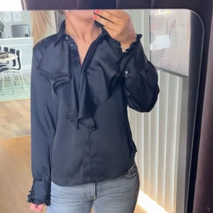 Volang blus ZARA - Säljer en svart satin blus med långa ärmar och stora volanger framtill och har knappar vid ärmsluten