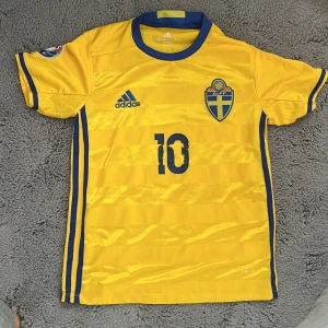 Sverige Zlatan Ibrahimović fotbollströja - Gul svensk landslagströja från Adidas med blå detaljer, nummer 10 och Ibrahimović på ryggen. Klassisk design med blå ränder på sidorna och landslagslogga på bröstet. Perfekt för dig som vill hylla Zlatan!