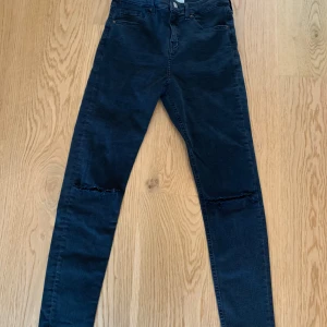 Svarta skinny jeans från Topshop Jamie - Svarta skinny jeans från Topshop Jamie med hög midja och slitna detaljer vid knäna. Klassisk femficksmodell och stretchigt material som sitter tight hela vägen ner. Perfekta till vardags för en cool look.