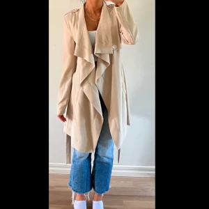 Beige trenchcoat med bälte - Säljer en beige trenchcoat med snyggt fall och knytbälte i midjan. Jackan har långa ärmar, bred krage och dekorativa knappar upptill. Perfekt för dig som vill ha en stilren och tidlös look.
