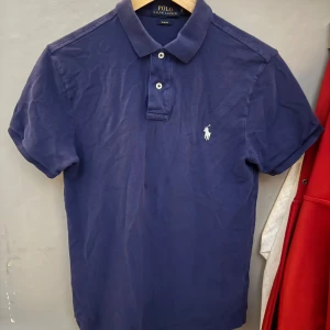 Blå piké t-shirt från Polo Ralph Lauren - Säljer en klassisk blå piké t-shirt från Polo Ralph Lauren med vit broderad logga på bröstet. Slim fit-modell med korta ärmar och två knappar vid kragen. Perfekt för en stilren och sportig look.