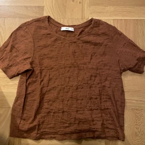 Brun kortärmad topp från MNG - Säljer en brun kortärmad topp från MNG i ett lätt, strukturerat tyg. Toppen har rund halsringning och en avslappnad passform, perfekt för varmare dagar.