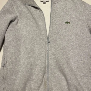 Grå ziptröja från Lacoste - Säljer en grå ziptröja från Lacoste med klassisk krokodillogga på bröstet. Tröjan har dragkedja framtill, två fickor och är tillverkad i mjukt material. Regular fit och långärmad modell, perfekt för en avslappnad stil.