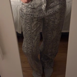 Gråa byxor med ormmönster - Säljer ett par gråa byxor med ormmönster och normal passform. Byxorna har hög midja och raka ben, perfekta för dig som vill sticka ut lite extra. Stängs med knappar och dragkedja framtill.