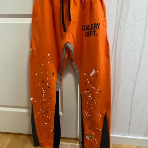 Gallery Dept byxor - Säljer ett par orange mjukisbyxor från Gallery Dept. med svart snörning i midjan och tryckt logga på ena benet. Byxorna har vita och svarta färgstänk samt svarta paneler längs benen. Mjukt material och bekväm passform. ❗️OBS PÅSEN MEDFÖLJER INTE❗️