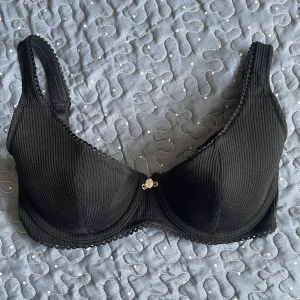 Svart ribbad bh från H&M med rosettdetalj - Svart bh från H&M med ribbad struktur och liten rosett framtill. Bh:n har vadderade kupor samt dekorativ kant längs kanterna. Perfekt för dig som vill ha en enkel men söt bh.