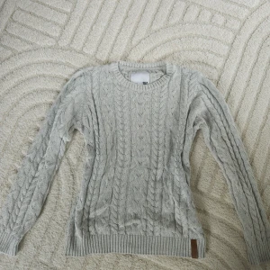 Ljusgrå stickad tröja med kabelmönster - Säljer en ljusgrå stickad tröja med klassiskt kabelstickat mönster. Tröjan har rund halsringning, långa ärmar och ribbade muddar vid ärmslut och nederkant. Perfekt för lager-på-lager under kyliga dagar.