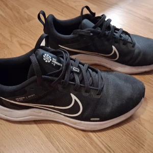 Svarta Nike sneakers med vit sula - Säljer ett par knappt använda svarta Nike Downshifter 12 sneakers med vit sula och vit swoosh på sidan. Skorna har snörning och en platt sula som ger bra grepp. Perfekta för träning eller vardagsbruk. Storlek US 8.5/UK 6 /EUR 40.  25,5 cm