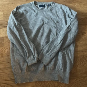Grå tröja från GANT - Säljer en klassisk grå tröja från GANT med rund halsringning och diskret broderad logga på bröstet. Tröjan har långa ärmar och ribbade muddar vid ärmslut och nederkant. Perfekt för en stilren vardagslook.